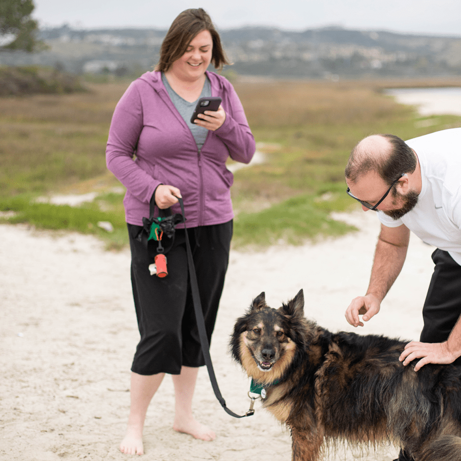 Larger_PetDesk_Case Studies_Increased Appointments_B Female pet parent utilizing her cell phone while holding leased dog and while male pet parent pets happy dog