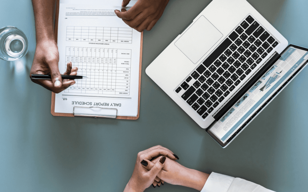 Person writing in a daily report schedule while chatting with another person with a laptop next to them