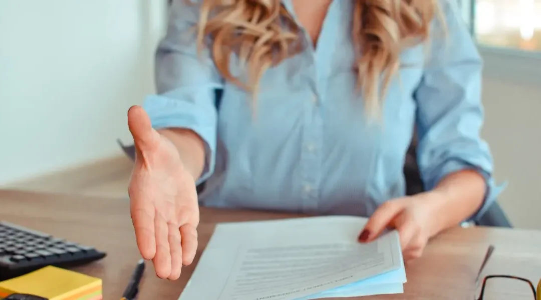 Women sitting at a desk while holding papers and holding her hand out for a handshake